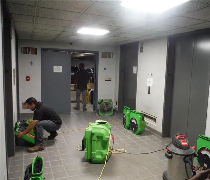 This parking corridor has two uniformed techs setting up bright green drying equipment after the elevators flooded